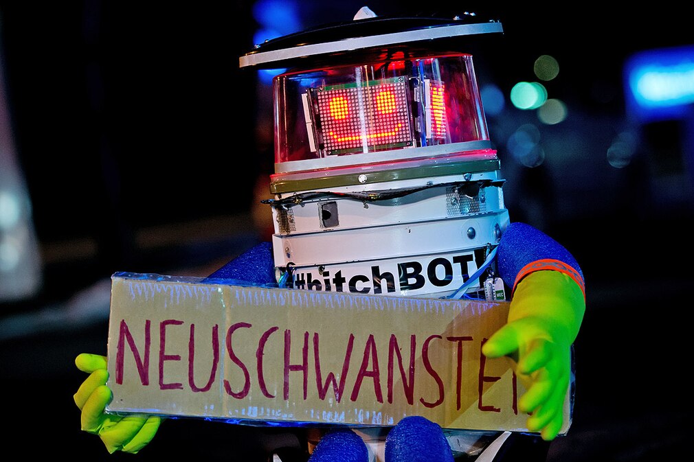 Der Roboter hitchBOT sitzt am 13.02.2015 in München mit einem Schild mit der Aufschrift Neuschwanstein am Straßenrand