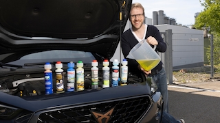 Redakteur steht neben Auto mit offener Motorhaube, dort sind die Scheibenreiniger-Flaschen aufgereiht. Redakteur gießt Reiniger in den Behälter vom Auto.