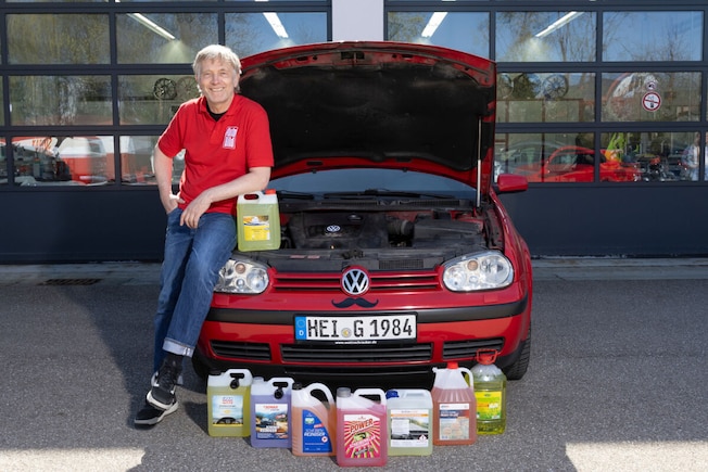 Redakteur steht neben Auto mit offener Motorhaube, dort sind die Scheibenreiniger-Flaschen aufgereiht. Redakteur gießt Reiniger in den Behälter vom Auto.