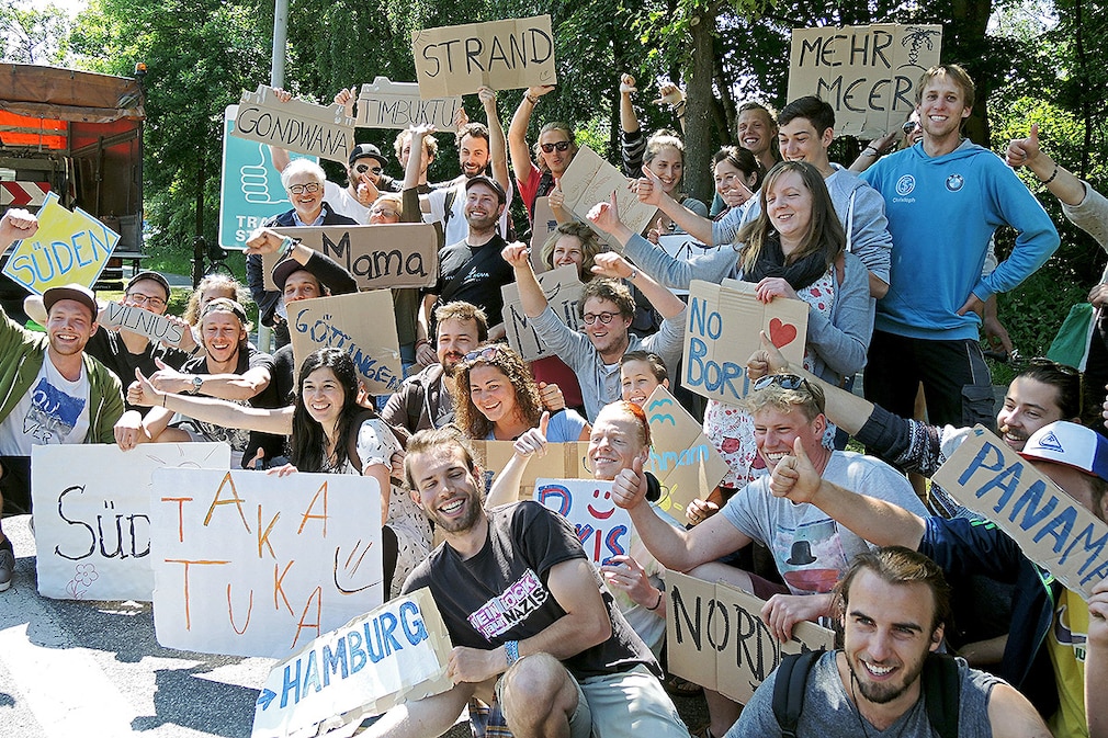 Die Initiatoren des Projekts, einige Tramper und der Bürgermeister der Stadt Kiel, Peter Todeskino (Bündnis 90/Die Grünen hinten links hält ein Schild mit der Aufschrift "Mama"), stehen am 11.06.2015 an einer sogenannten «Trampstelle» in Kiel (Schleswig-Holstein). Die nach Angaben der Initiatoren deutschlandweit erste offizielle Haltestelle für Rucksacktouristen ist am 11.06.2015 in Kiel eröffnet worden. Foto: Claudia Thaler/dpa
