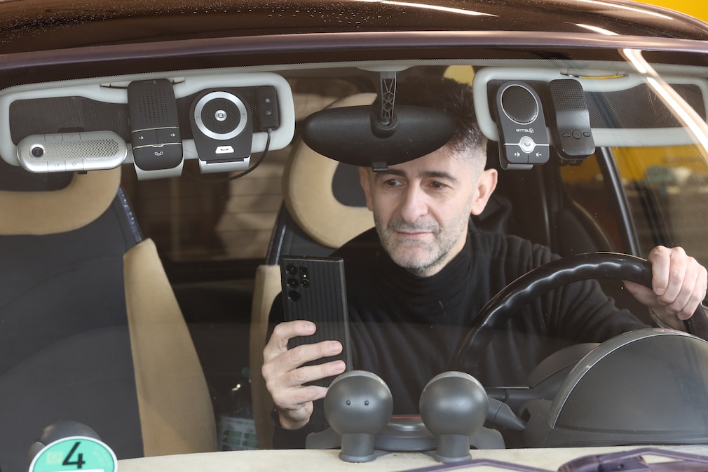 Symbolfoto: Eine Person sitzt mit Handy in der Hand im Auto, umgeben von im Cockpit befestigten Freisprechanlagen
