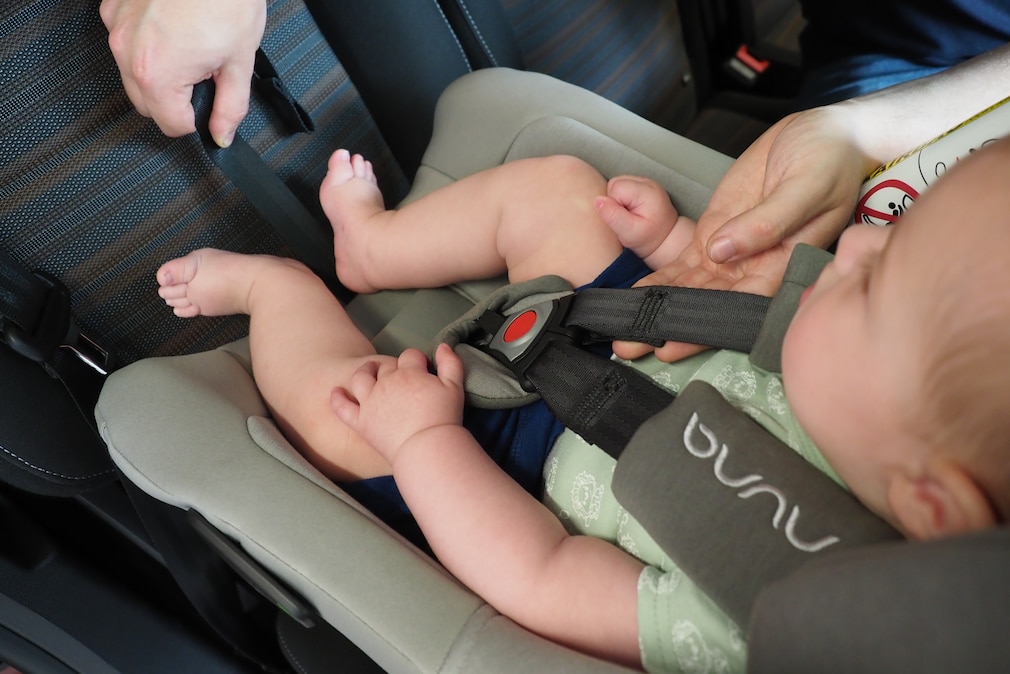 Babyschale mit Baby: Eine Hand zeigt, wie sich der Gurtstraffer bedienen lässt.