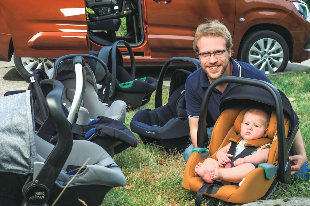 Eine Person hockt hinter einer Babyschale mit Baby. Umgeben von allen weiteren getesteten Babyschalen Im Hintergrund ist ein Auto mit geöffneter Schiebetür zu erkennen.