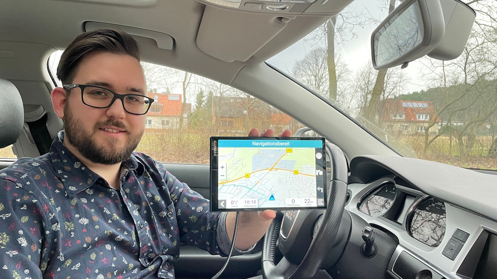 Eine Person sitzt in einem Fahrzeug und hält dem Betrachter eine Navi-Display mit abgebildeter Karte entgegen