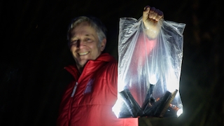 Eine Person hält bei Dunkelheit eine Klarsichtplatiktüte mit vielen leuchtenden Taschenlampen in die Höhe.