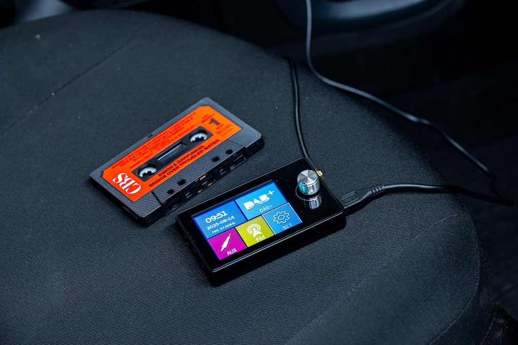 Größenvergleich: Ein DAB+-Adapter liegend auf dem Autositz neben einer Musikkassette