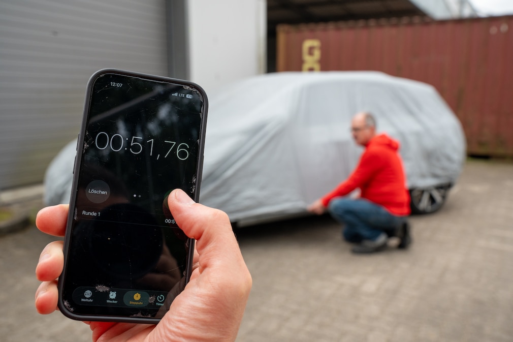 Im Vordergrund ein Smartphone mit laufender Stoppuhr, im Hintergrund eine Person beim Befestigen einer Abdeckplane an einem VW Golf.