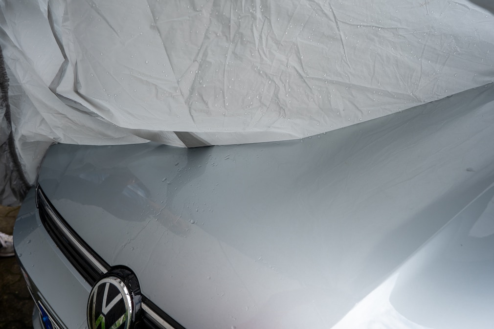 Nahansicht einer von der Motorhaube gehobenen Plane. Auf dem Fahrzeug sieht man deutlicher Wasserspuren im Nahtbereich.