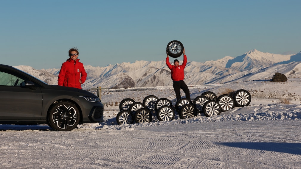 Redakteure stehen vor einer Reihe Reifen, ein Redakteur hebt einen Reifen an. An der Seite steht ein grauer VW Golf