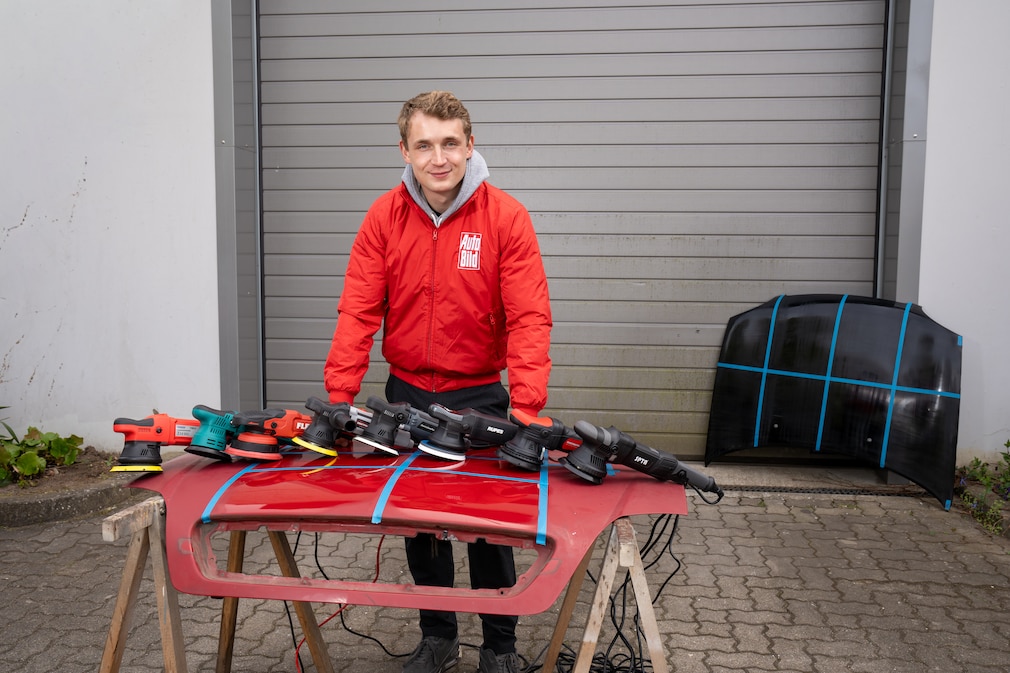 Eine Person steht vor einer in Felder eingeteilten Motorhaube auf der 8 Exzenter-Poliermaschinen liegen.