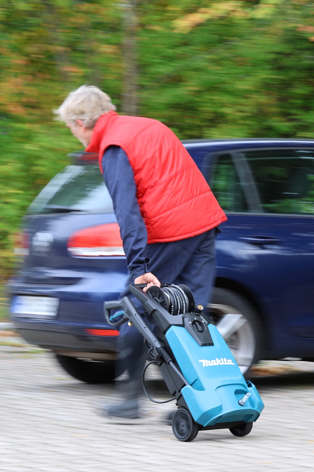 Person zieht in gebeugtem Gang einen Hochdruckreiniger mit zu kurzem Transportgriff hinter sich her.