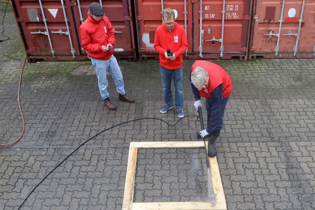 Testsituation: 3 Pesrsonen bei Lautstäke- und Zeitmessung. Eine Person arbeitet mit Hochdruckreiniger am festgelegten Normquadratmeter, definiert durch einen Holzrahmen.