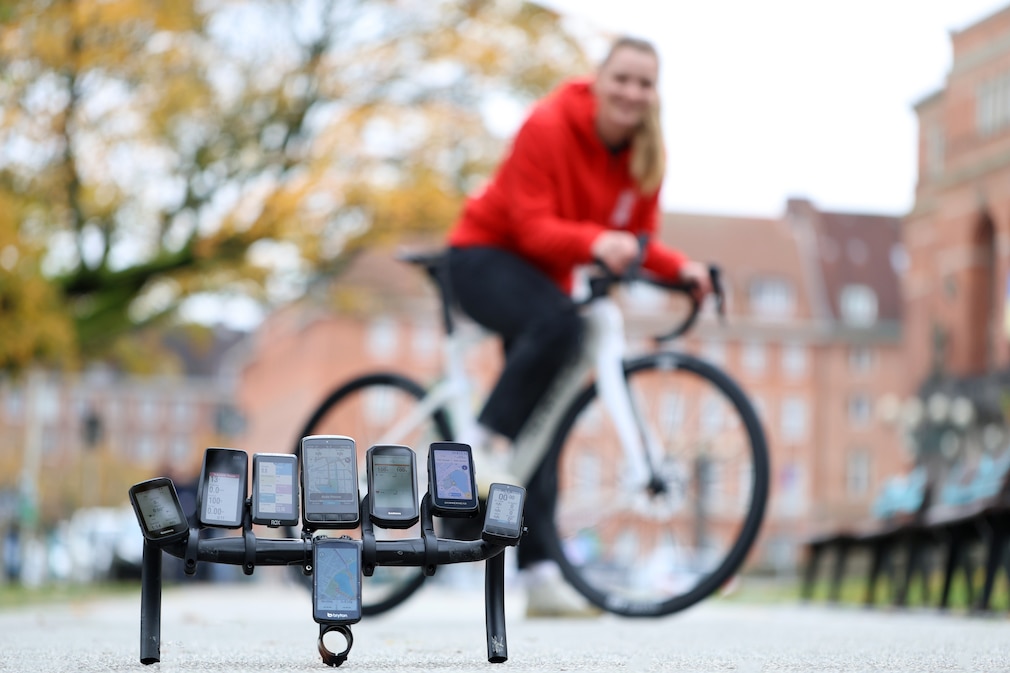 Straßensituation: Ein Rennradlenker mit acht montierten Fahrradcomputern liegt auf der Straße. Im Hintergrund steht eine Person mit einem Rennrad.