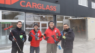 Vier Personen, mit Eiskratzern in der Hand, stehen auf einer Fläche vor einem geöffneten Tiefkühlcontainer, in dem sich eingefrorene Autoscheiben befinden.