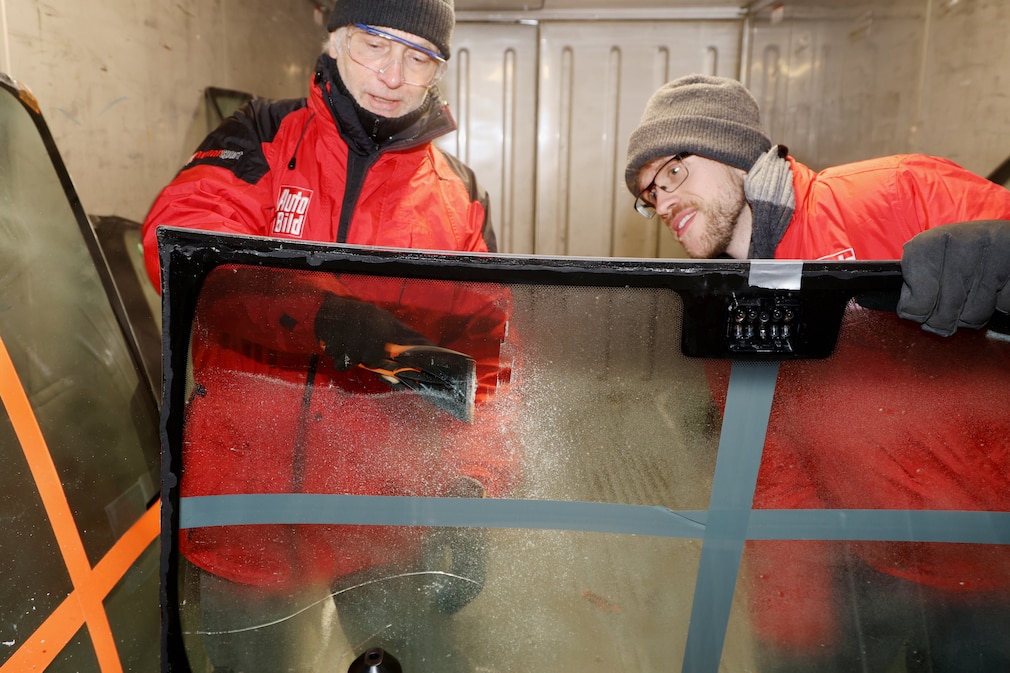 Zwei Männer kratzen Eis in einem Kühlcontainer auf einer ausgebauten Frontscheibe