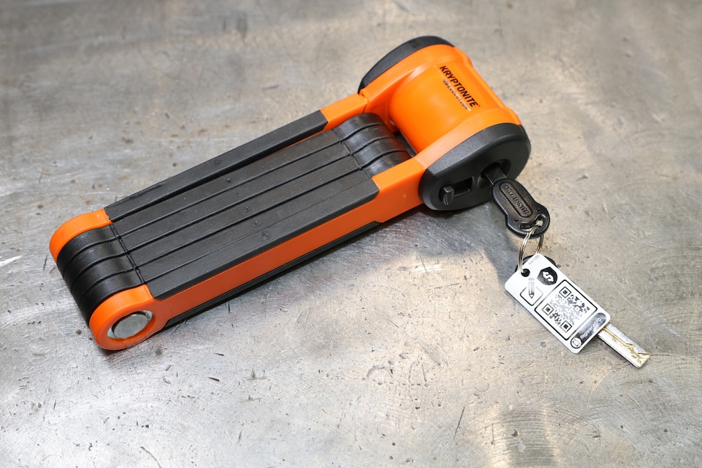 Produktaufnahme des orange-schwarzen Kryptonite Faltschlosses auf einem Werktisch aus Stahl