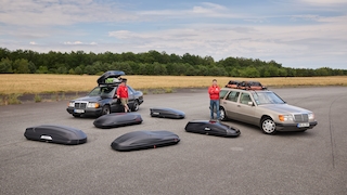 Dachboxen liegen verteilt auf einem Asphalt, dahinter zwei Redakteure in roten AUTO BILD-Jacken, die an jeweils einem Mercedes W124 lehnen, die jeweils eine Dachbox auf dem Dach haben.