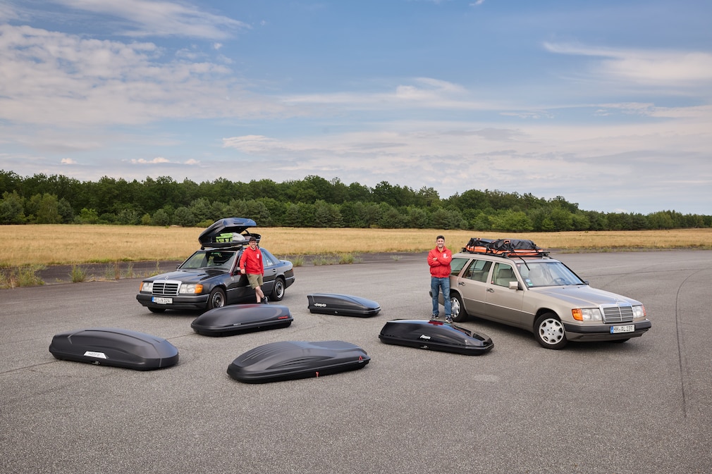 Dachboxen liegen verteilt auf einem Asphalt, dahinter zwei Redakteure in roten AUTO BILD-Jacken, die an jeweils einem Mercedes W124 lehnen, die jeweils eine Dachbox auf dem Dach haben.