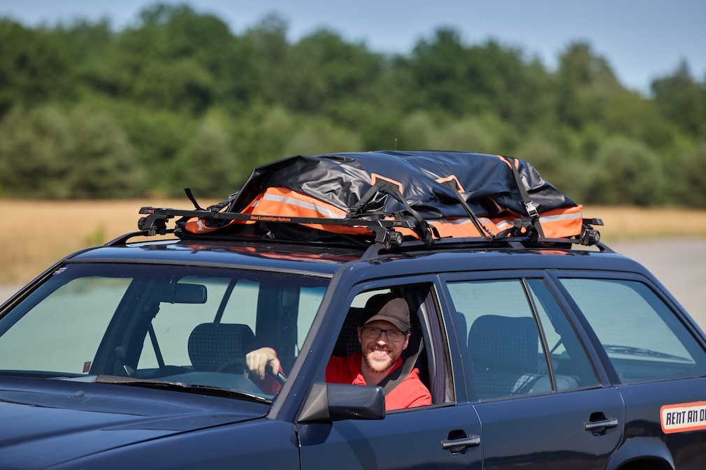 Redakteur lehnt aus dem Fahrerfenster eines blauen Mercedes, auf dem Dach eine Dachbox.