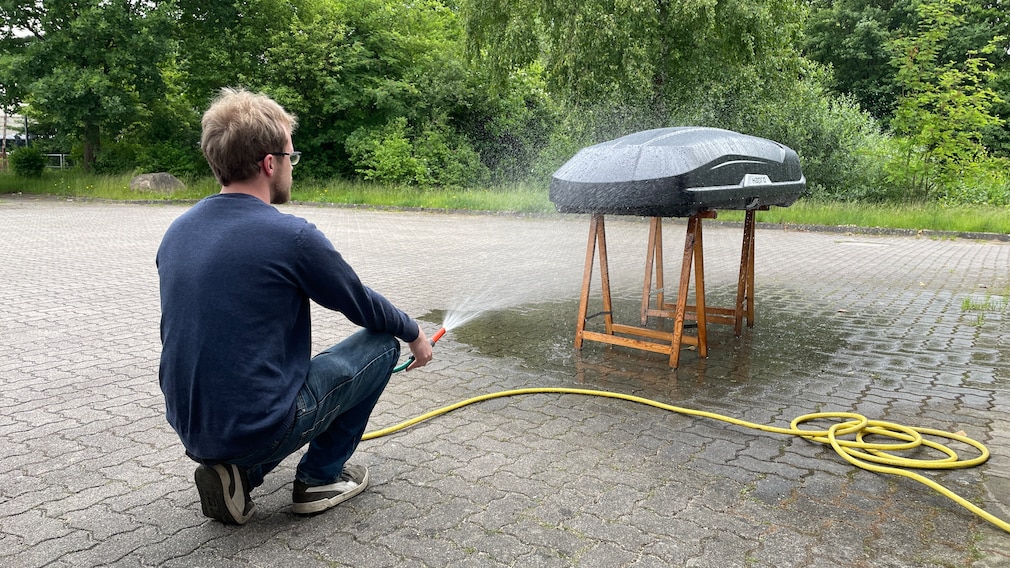 Testsituation: Eine Person besprüht eine aufgebockte Dachbox mit Wasser, um die Dichtigkeit zu testen