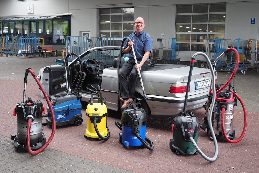 Eine Person, sitzend auf einem geöffneten Cabriolet, davor stehend 6 Nass-Trockensauger.