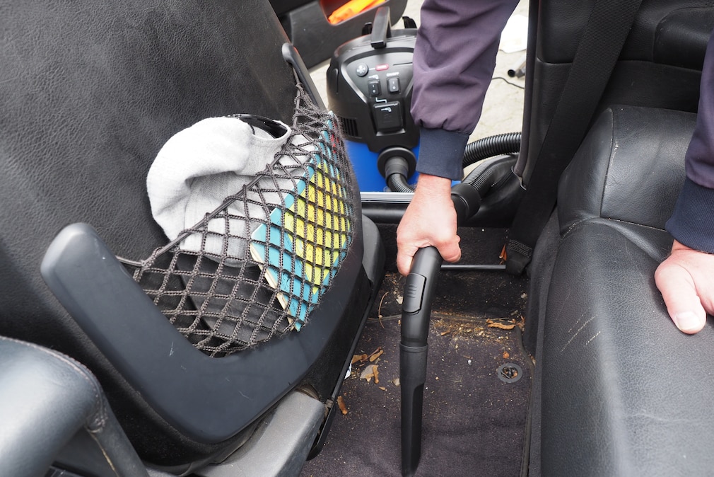 Person saugt verschmutzten Fahrzeugteppich vor der Rücksitzbank eines Fahrzeugs.
