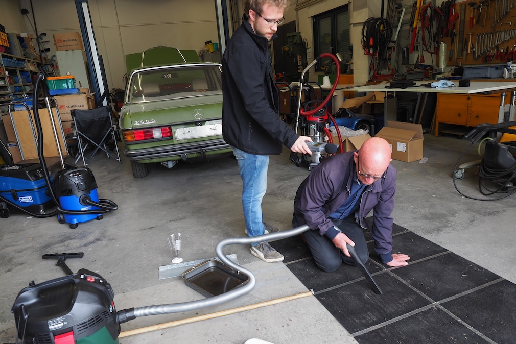 Werkstattsituation: eine Person arbeitet mit einem Sauger, kniend auf einer in mehrere Flächen unterteilten Versuchsfläche. Eine zweite Person steht mit einem Phon-Messgerät daneben.