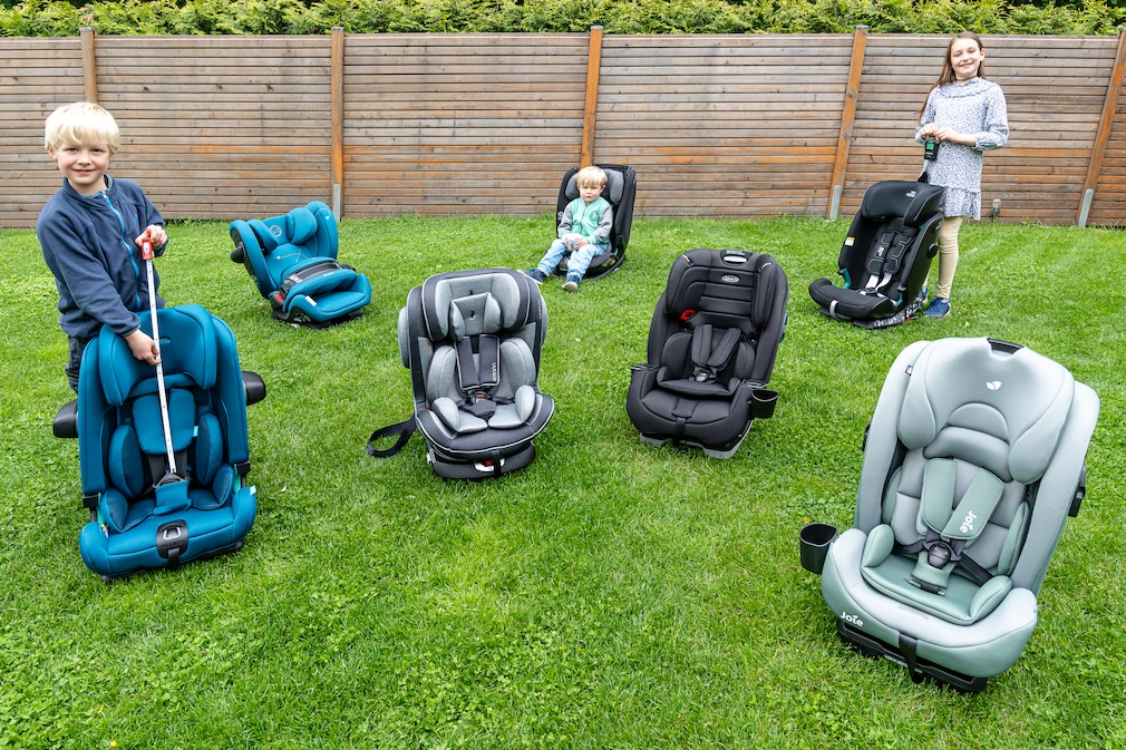 Drei unterschiedlich große Kinder stehen und sitzen verteilt zwischen sieben Kindersitzen auf einer Grünfläche.