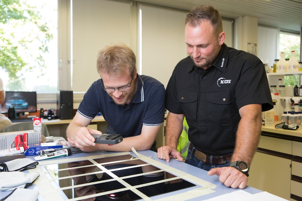 Laborsituation: zwei Personen betrachten mit einem Analysegerät das Lackbild der Testflächen auf einem Arbeitstisch.