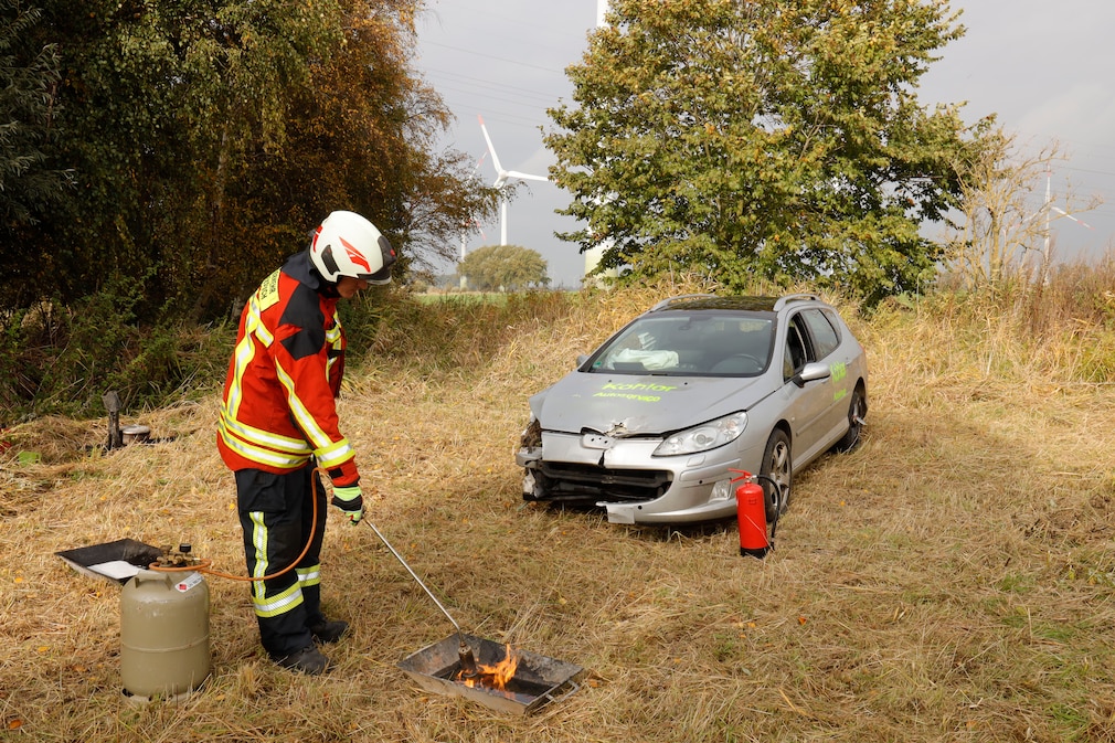 Testaufbau auf einer Weide: Ein Feuerwehrmann versucht eine Flüssigkeit in einer Wanne zu entzünden. Dahinter steht ein Unfallfahrzeug.