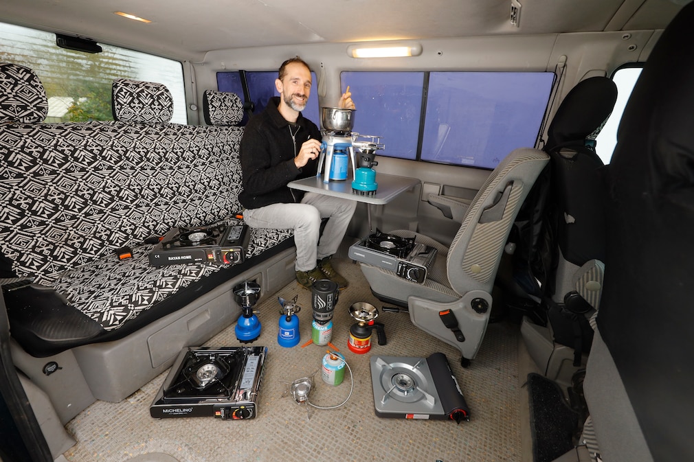 Eine Person sitzt in einem VW-Bus auf der Rücksitzbank am Tisch und ist von verschiedenen, um sie herum arrangierten Campingkochern umgeben.
