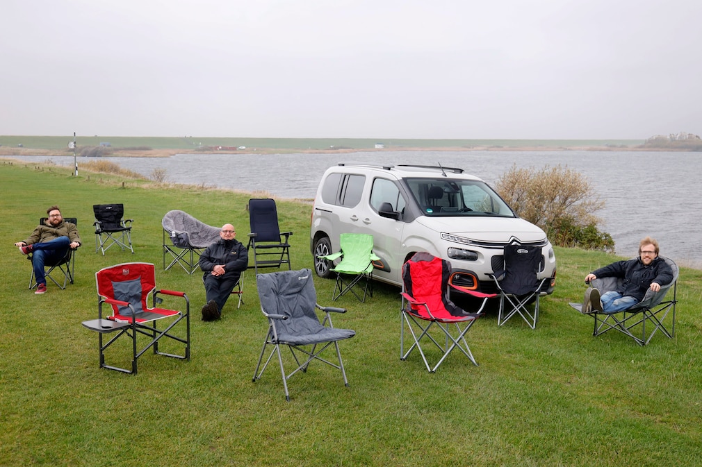 Drei Personen sitzen auf Campingstühlen, umgeben von weiteren Campingstühlen auf einer Wiese vor einem Auto an einer Wasserfläche.