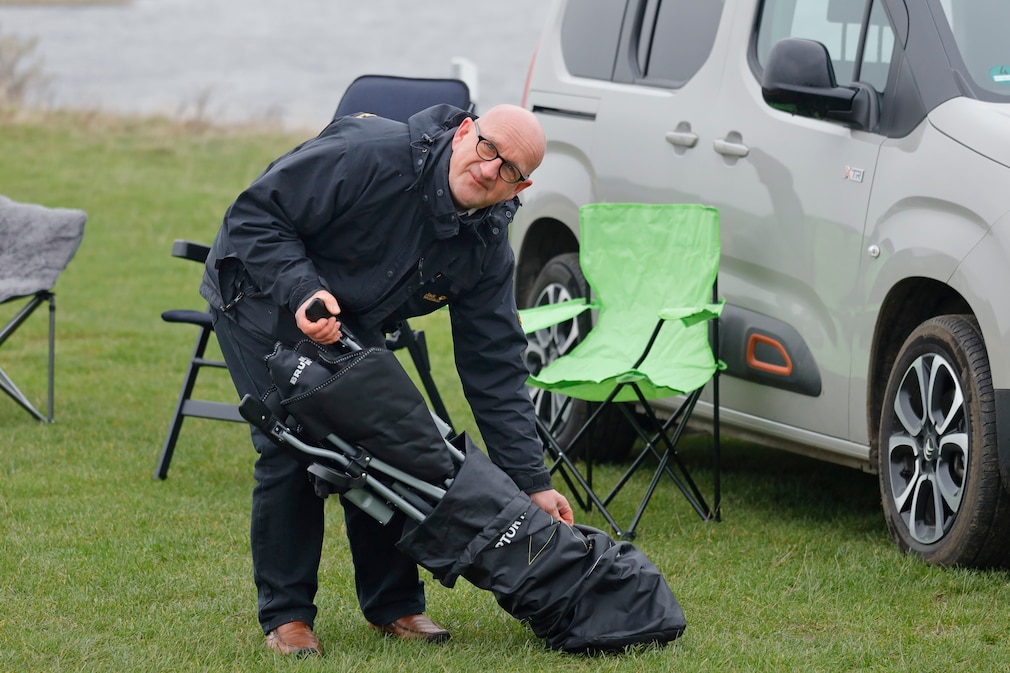 Eine Person zieht einen Campingstuhl aus der Transporthülle, stehend auf einer Wiese vor einem Auto