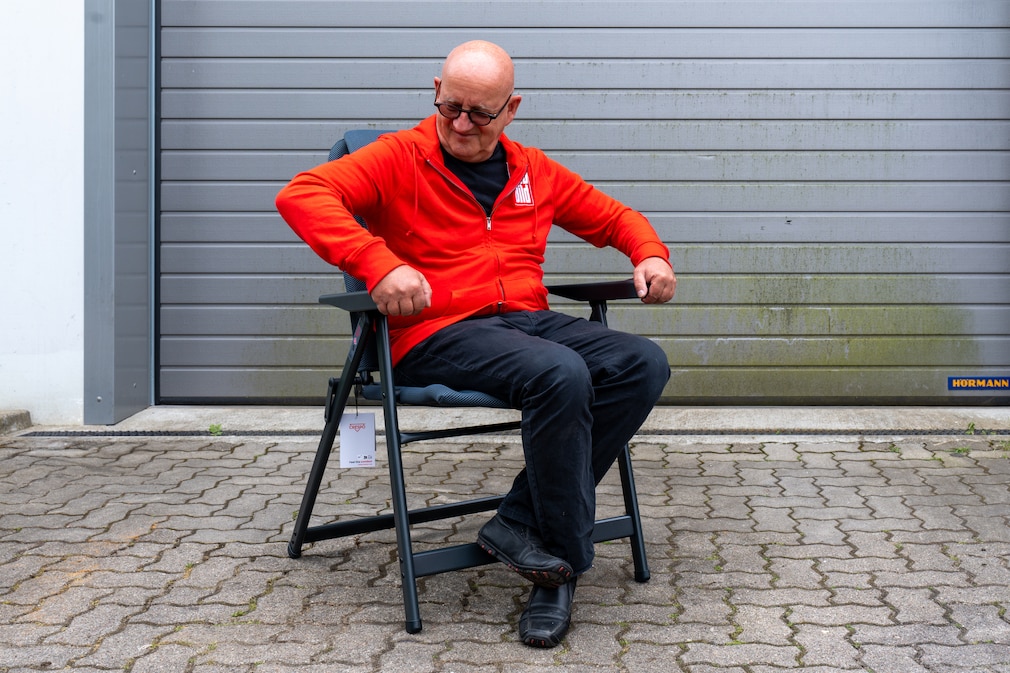 Nahaufnahme einer Person, die das Gestell eines zerlegten Campingstuhls hält. Im Hintergrund eine geöffnete Heckklappe