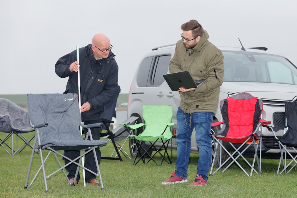 Testsituation: Zwei Personen vermessen und beschreiben Campingstühle auf einer Wiese, stehend vor einem Auto