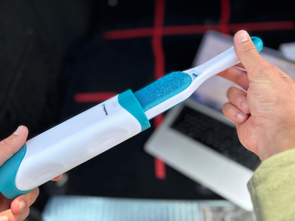 Nahaufnahme: Produktfoto eines Tierhaarentferners, der grad aus seiner Aufbewahrungshülle gezogen wird. Unscharf dahinter weitere Testprodukte, liegend auf der Testfläche.