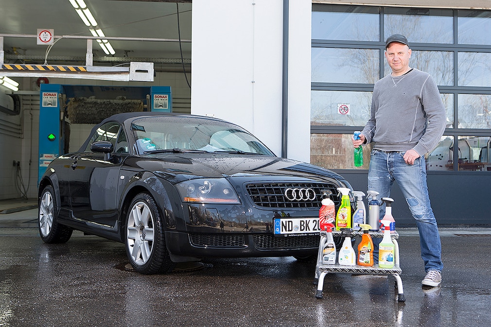 Eine Person steht neben einem Fahrzeug vor einer Waschanlage. Im Vordergrund sind Insektenentferner in zwei Reihen auf einem Tritt aufgereiht.