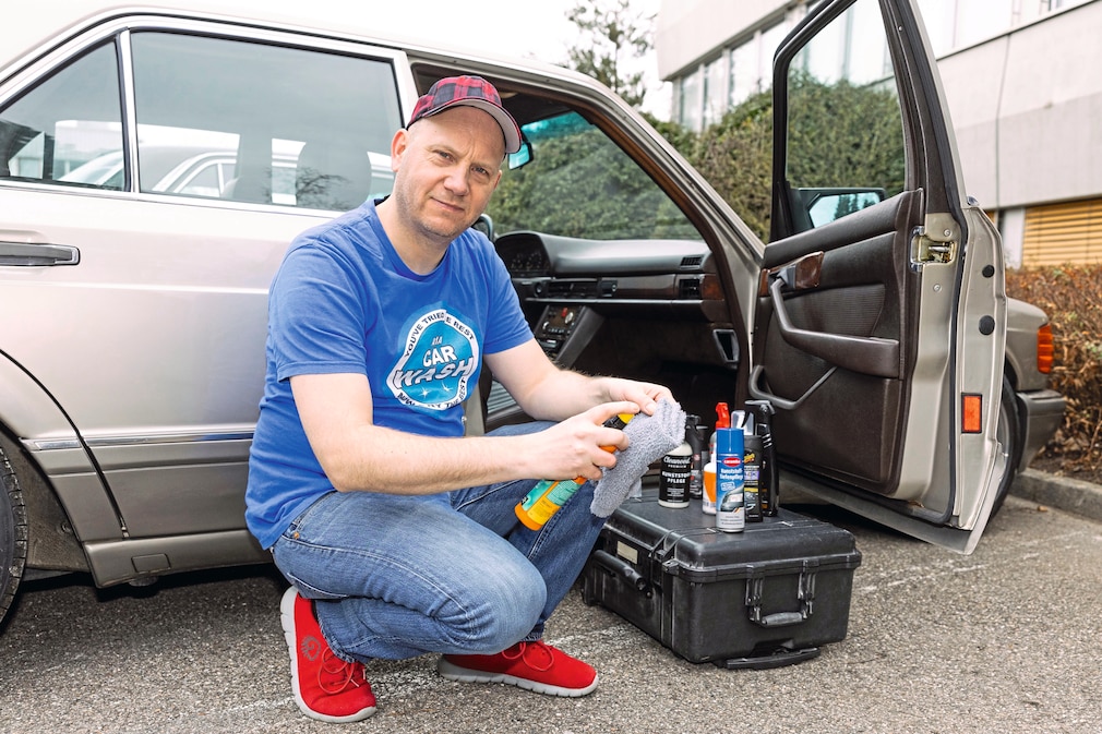 Eine Person kniet mitt einem Lappen vor einer offenen Fahrzeugtür, davor aufgereiht auf einem Koffer verschiedene Kunststoffpfleger-Tests.