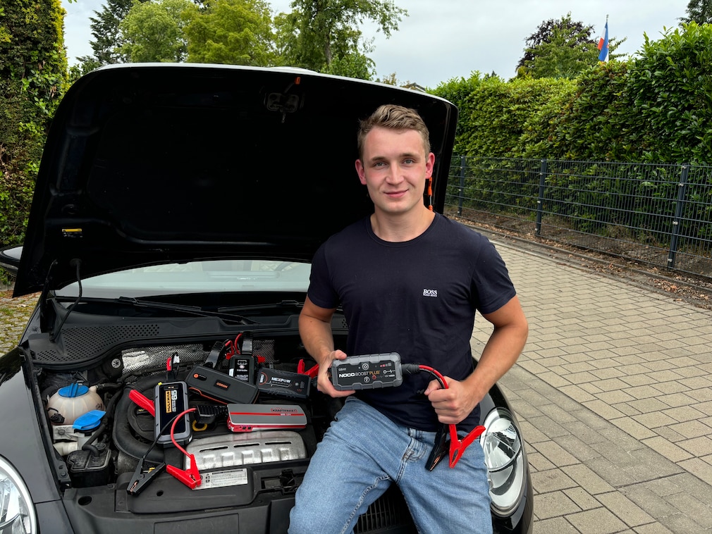 Person sitzt vor geöffneter Motorhaube am Motorraum, mit einem Starthilfegerät in der Hand, alle weiteren getesteten Geräte sind zur Demonstration im Motorraum verteilt.