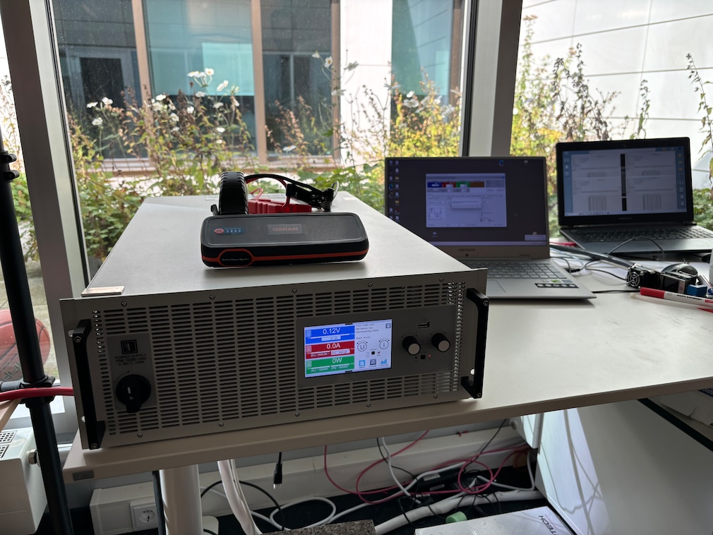 Starthilfegerät liegt zu Demo-Zwecken auf dem Power Supply PSB Netzgerät welches auf einem Schreibtisch steht, im Hintergrund 2 Laptops.