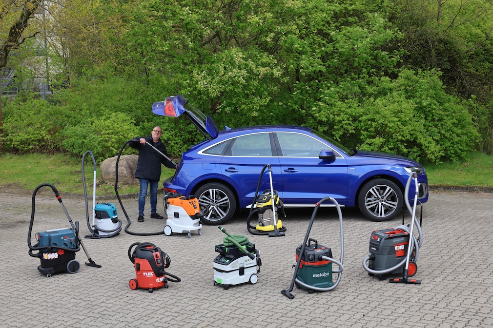Seitenansicht: Person steht mit einem Staubsauger arbeitend an einer geöffneten Fahrzeugheckklappe, alle Testprodukte stehen verteilt vor dieser Szene.