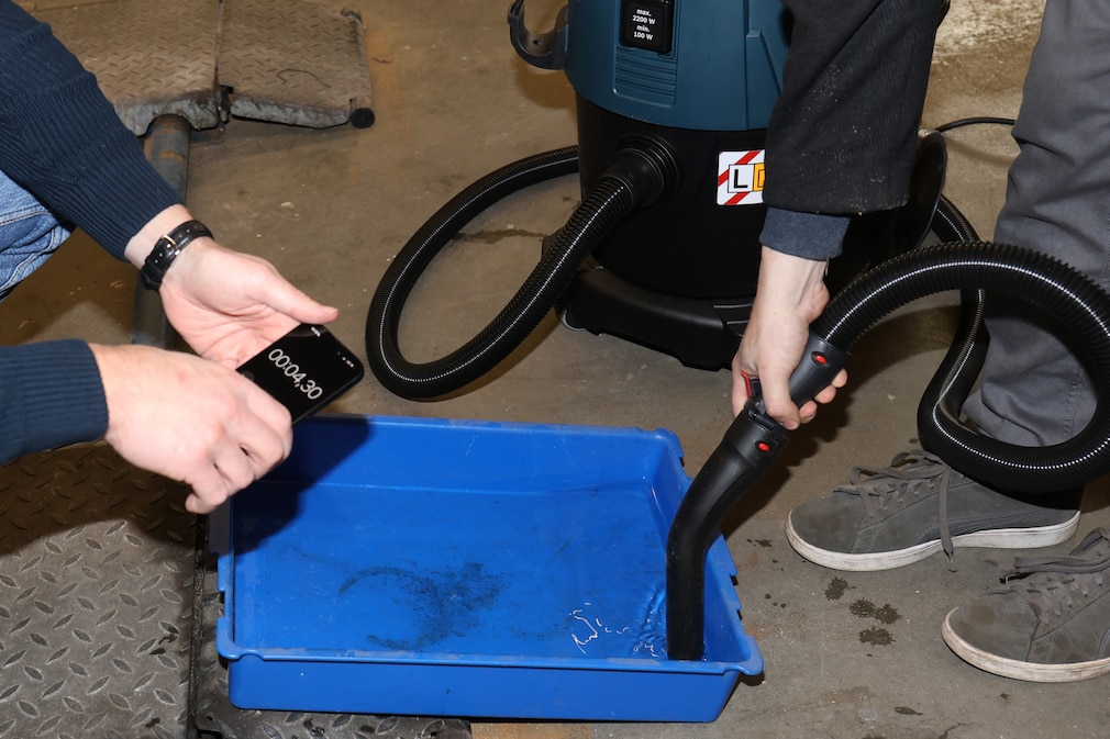 Testsituation: Nahaufnahme von Zeitnahme bei Ermittlung der Nasssaugleistung während mittels einer Staubsaugerdüse Wasser aus einer Plastikwanne gesaugt wird.