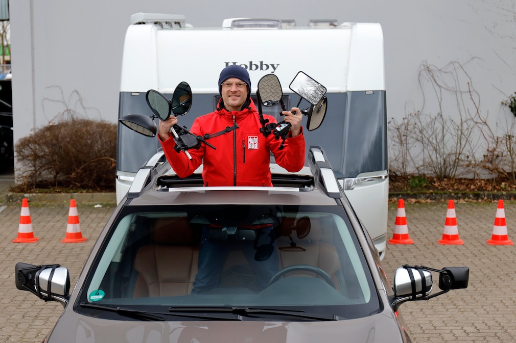 Standfoto: eine Person steht im offenen Schiebedach eines Fahrzeugs und hält verschiedene Zusatzspiegel in den Händen. Hinter dem Fahrzeug angehängt steht ein Wohnwagen.