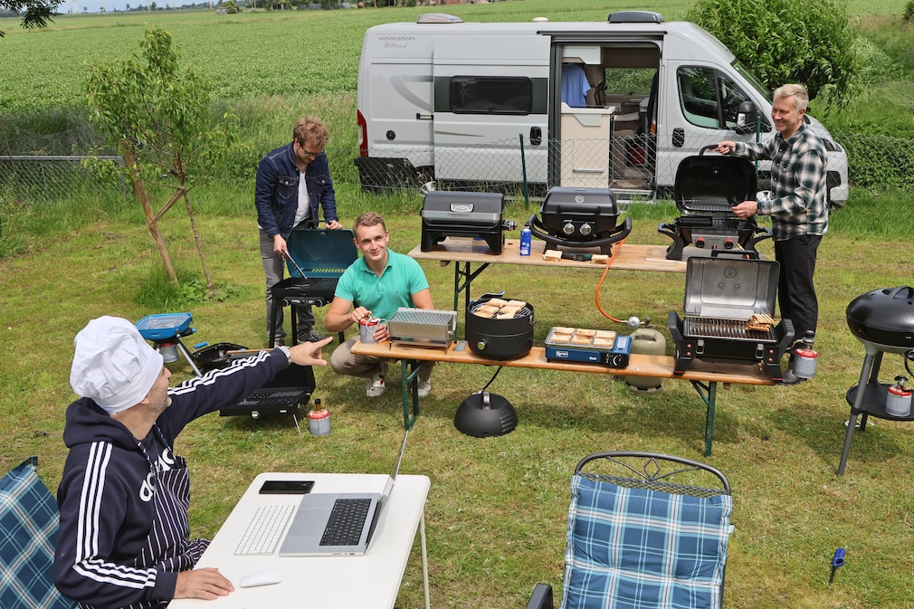 Vier Personen stehen auf einer Rasenfläche und bewerten Camping-Gasgrills. Im Hintergrund steht ein geöffnetes Wohnmobil.