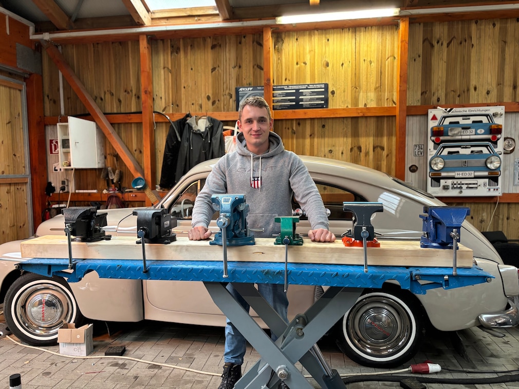 Werkstattsituation: eine Person steht hinter sechs auf einer Ebene aufgereihten Schraubstöcken. Hinter der Szene steht ein Fahrzeug.