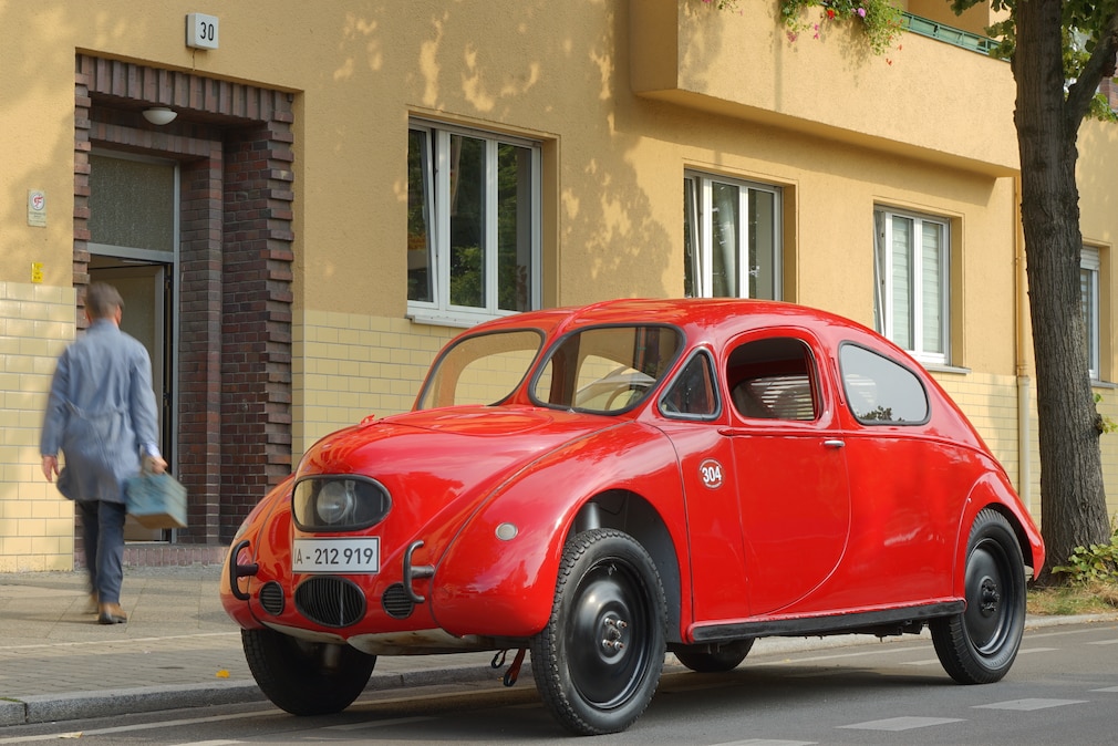 Das Auto steht vor einem gelben Haus, ein Mann im grauen Kittel geht hinein.