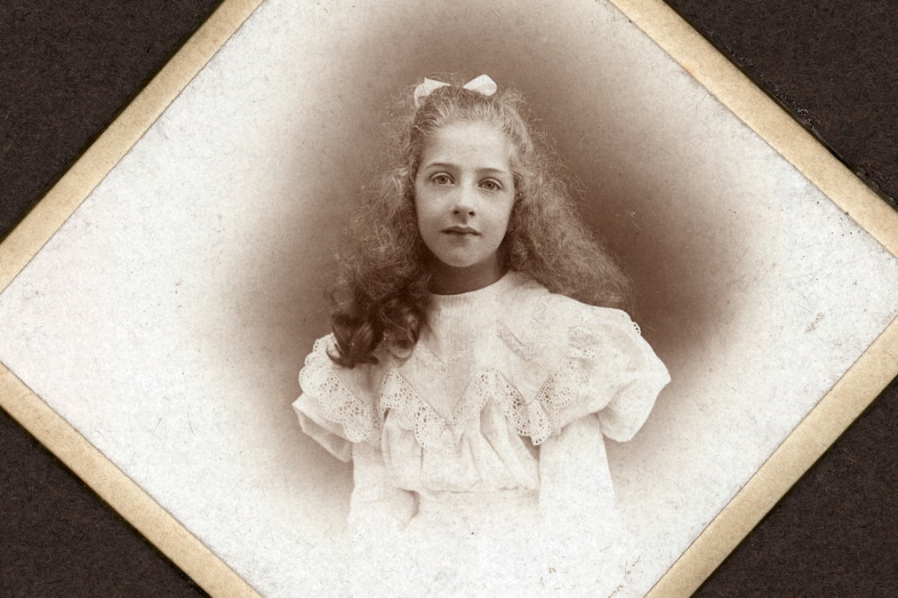 Etwa zwölfjähriges Mädchen mit Schleife im lockigen Haar, helles Kleid.