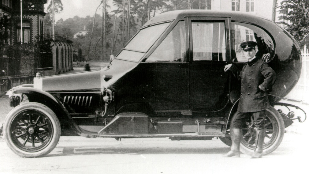 Automobil von 1912 mit einer Karosserie, die hinten rund ist wie ein Ei. Davor steht ein Mann in Uniform. Historisches Foto.