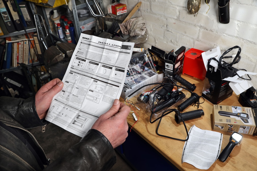 Werkstattsituation: eine Person im Anschnitt ließt eine Montage- und Betriebsanleitung, im Hintergrund diverse ausgepackte Motorradheizgriffe auf einer Arbeitsplatte.