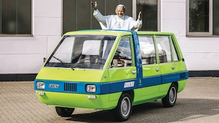 Grün-blauer Minitbus von schräg vorn; Papst Johannes Paul II. als Papp-Aufsteller schaut aus dem offenen Dach heraus